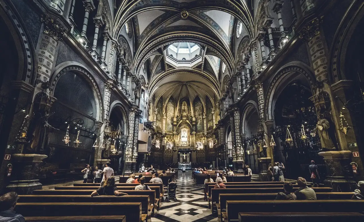 The arched, gilded nave of the Montserrat basilica, with rows of wooden pews and a few visitors
