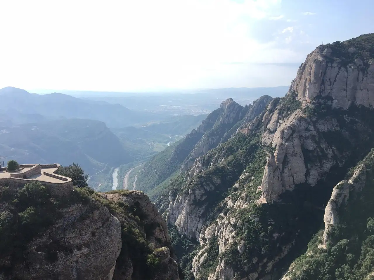 The jagged rock peaks of Montserrat above a deep green valley, with a stone viewpoint terrace on the left