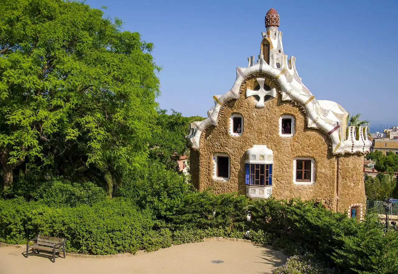 A Gaudí gatehouse at Park Güell with a wavy tiled roof and a tower, among green trees