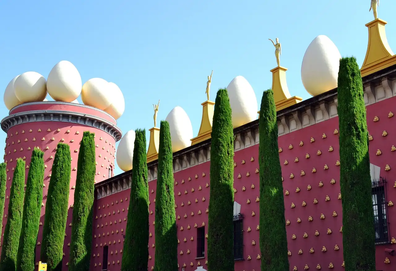 The red wall of the Dalí Theatre-Museum in Figueres, topped with giant white eggs, behind tall cypress trees