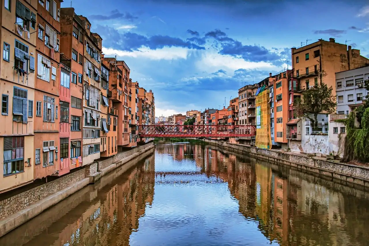 Tall painted houses reflected in the Onyar river in Girona, with a red iron bridge