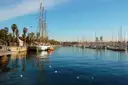A tall wooden sailing ship moored by the palms of Port Vell, with Barcelona's marina behind
