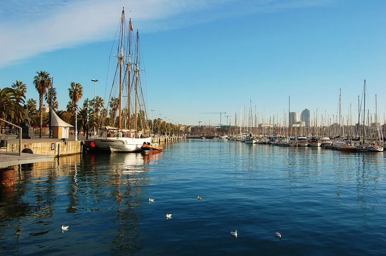 A tall wooden sailing ship moored by the palms of Port Vell, with Barcelona's marina behind