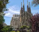 The spires of the Sagrada Família rising behind green and red trees under a blue sky