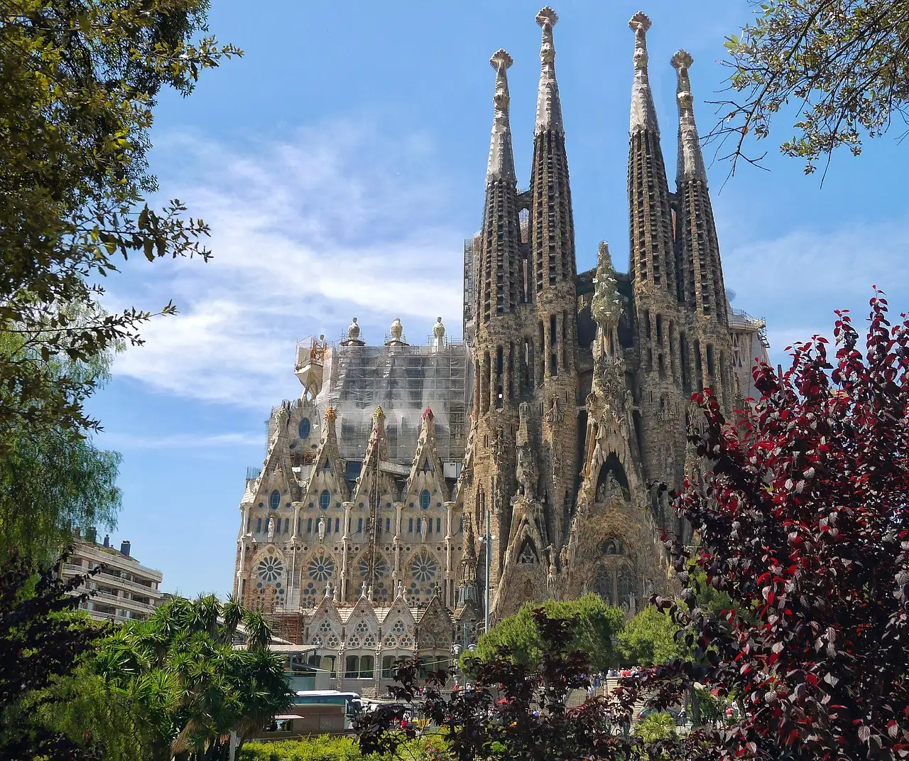 The spires of the Sagrada Família rising behind green and red trees under a blue sky